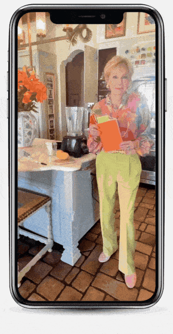 A woman in colorful clothes stands in a kitchen holding an orange book, seen through a phone screen by a counter with flowers and blender.
