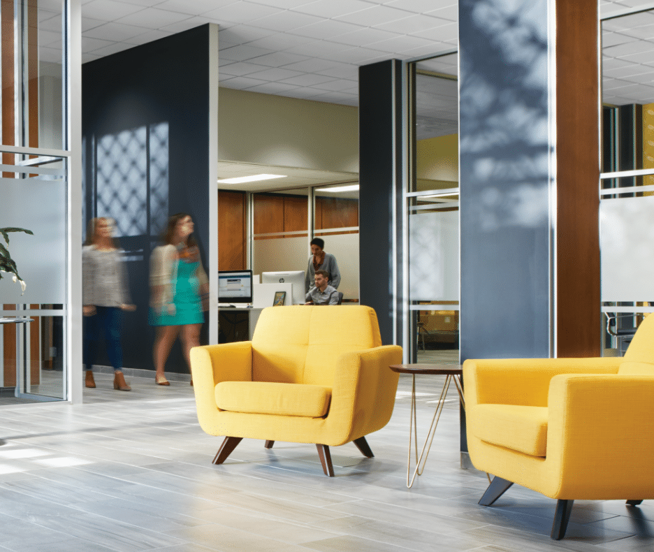 Modern office featuring two yellow armchairs in the foreground, glass walls, and three people walking in the background. One person sits at a desk as sunlight casts patterns on the walls and floor.